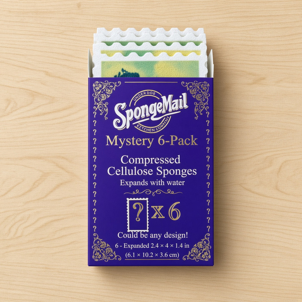 A purple box of SpongeMail Mystery 6-Pack compressed cellulose sponges, shown open on a light wood background. The box features gold decorative borders and text, with several colorful sponges visible at the top.