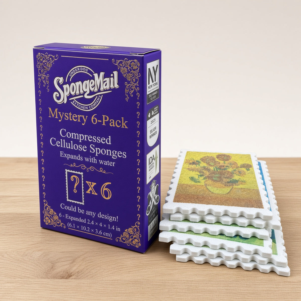 A purple SpongeMail Mystery 6-Pack box stands on a light wood surface next to a stack of white postage-stamp-shaped compressed cellulose sponges, one of which is expanded to show a sunflower design.